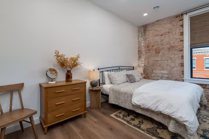 Bedroom with Brick Wall and Iron Bed Frame Bedroom with brick accent wall, black iron bed frame, neutral patterned bedding, and wood dresser with dried florals