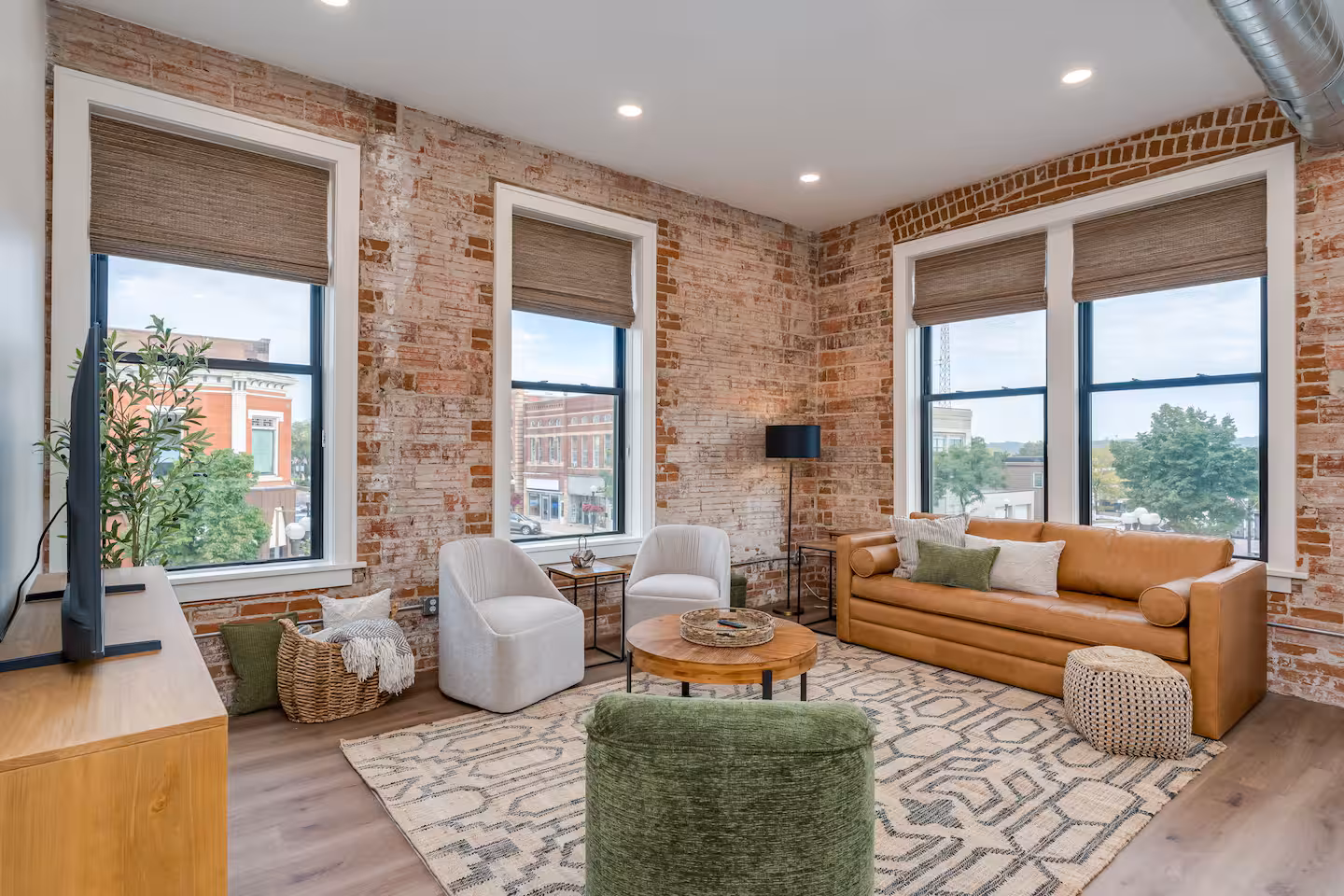 Spacious living room at Guten Tag Lofts featuring historic exposed brick walls, tall windows, a tan leather sofa, and modern white armchairs.