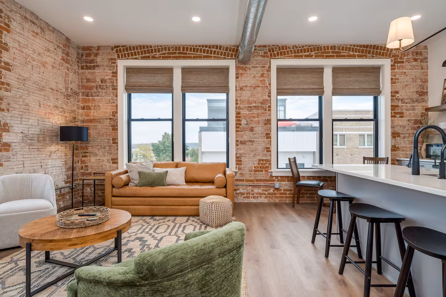 Living Room and Kitchen with Exposed Brick Wall Open-concept living room and kitchen with exposed brick wall, tan leather sofa, olive green armchair, and large windows overlooking downtown New Ulm